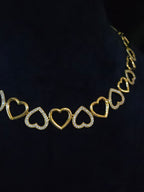 A gold heart-link necklace displayed on a black velvet jewelry bust. The necklace features alternating solid gold heart links and open heart links paved with small clear stones.