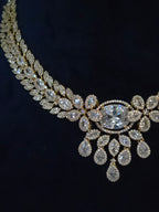 An ornate gold-toned necklace with multiple rows of clear teardrop and oval gemstones, featuring a large central round stone, displayed against a dark navy background.