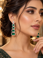 A close-up of a woman wearing a long drop earring with four rectangular green emerald-cut stones in a silver-toned setting, with a matching earring visible on the other ear. The background is a softly blurred warm indoor setting.