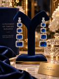 A pair of long drop earrings with rectangular blue gemstones and clear crystal-like borders, displayed on a dark blue velvet jewelry stand. A matching blue jewelry box is visible behind, with a marble surface and elegant decorative background.