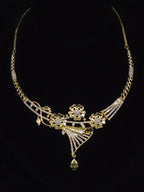 Gold-toned statement necklace with floral motifs, pavé-style clear stones, and a dangling bell-shaped pendant, displayed against a black background.