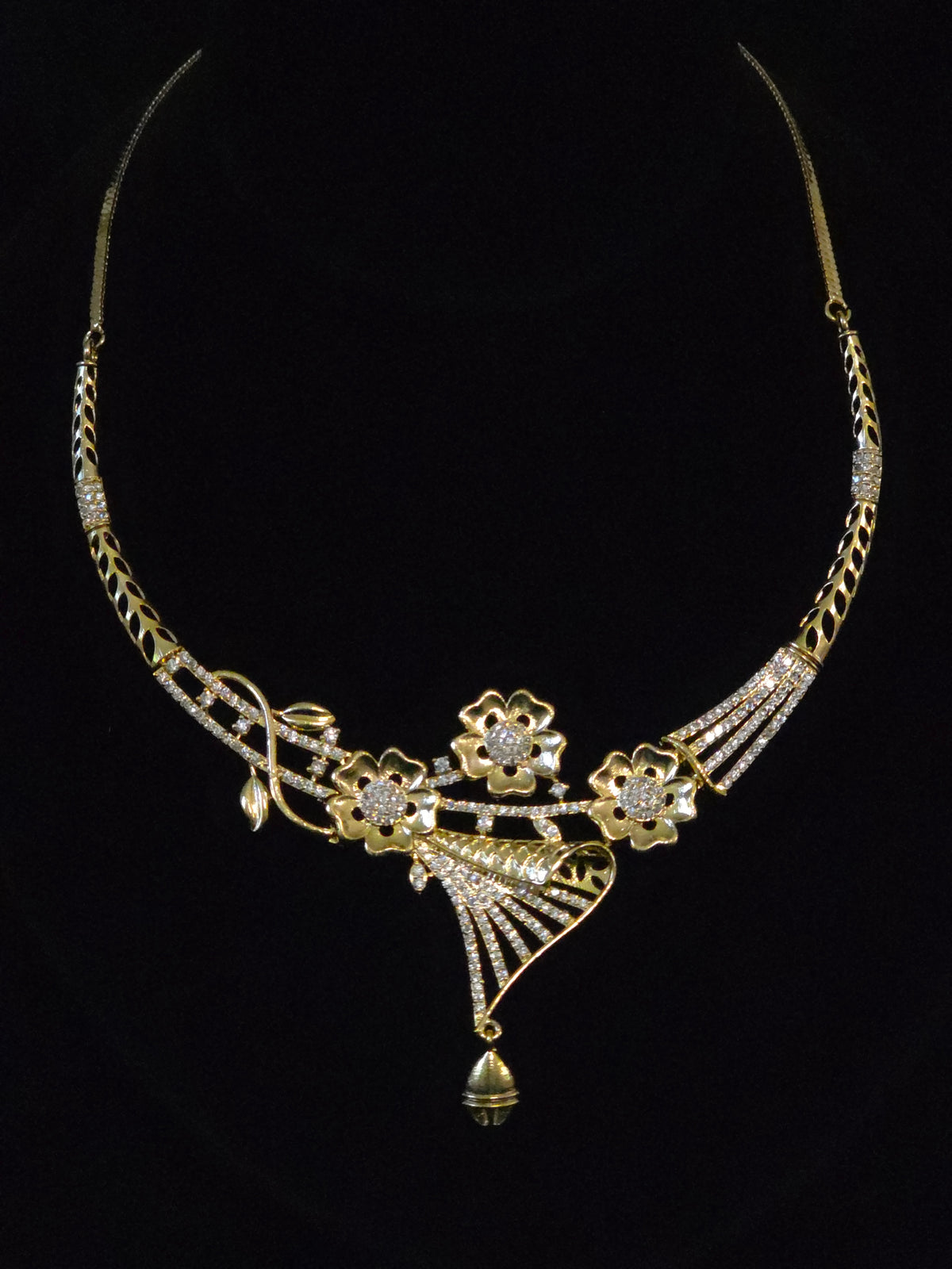 Gold-toned statement necklace with floral motifs, pavé-style clear stones, and a dangling bell-shaped pendant, displayed against a black background.
