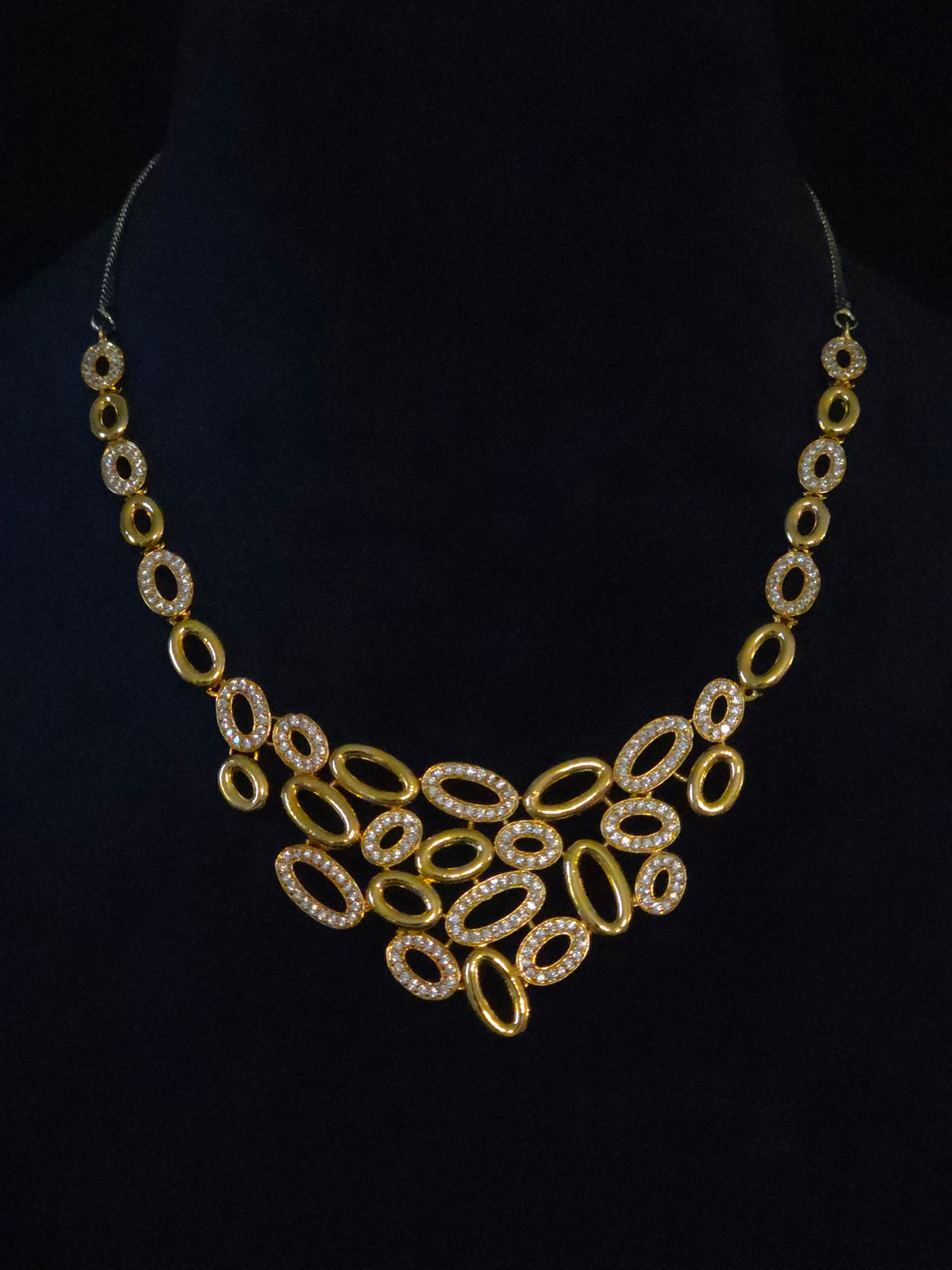 A gold statement necklace with a bib-style arrangement of oval links, some encrusted with clear rhinestones, displayed on a black mannequin bust.