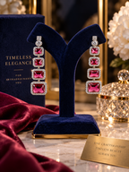 A pair of long drop earrings with rectangular pink gemstones and silver-toned crystal borders, displayed on a dark blue velvet jewelry stand. A matching dark blue box labeled 'TIMELESS ELEGANCY' sits behind the stand, with a marble surface, draped burgundy fabric, and a decorative gold-toned container in the background.
