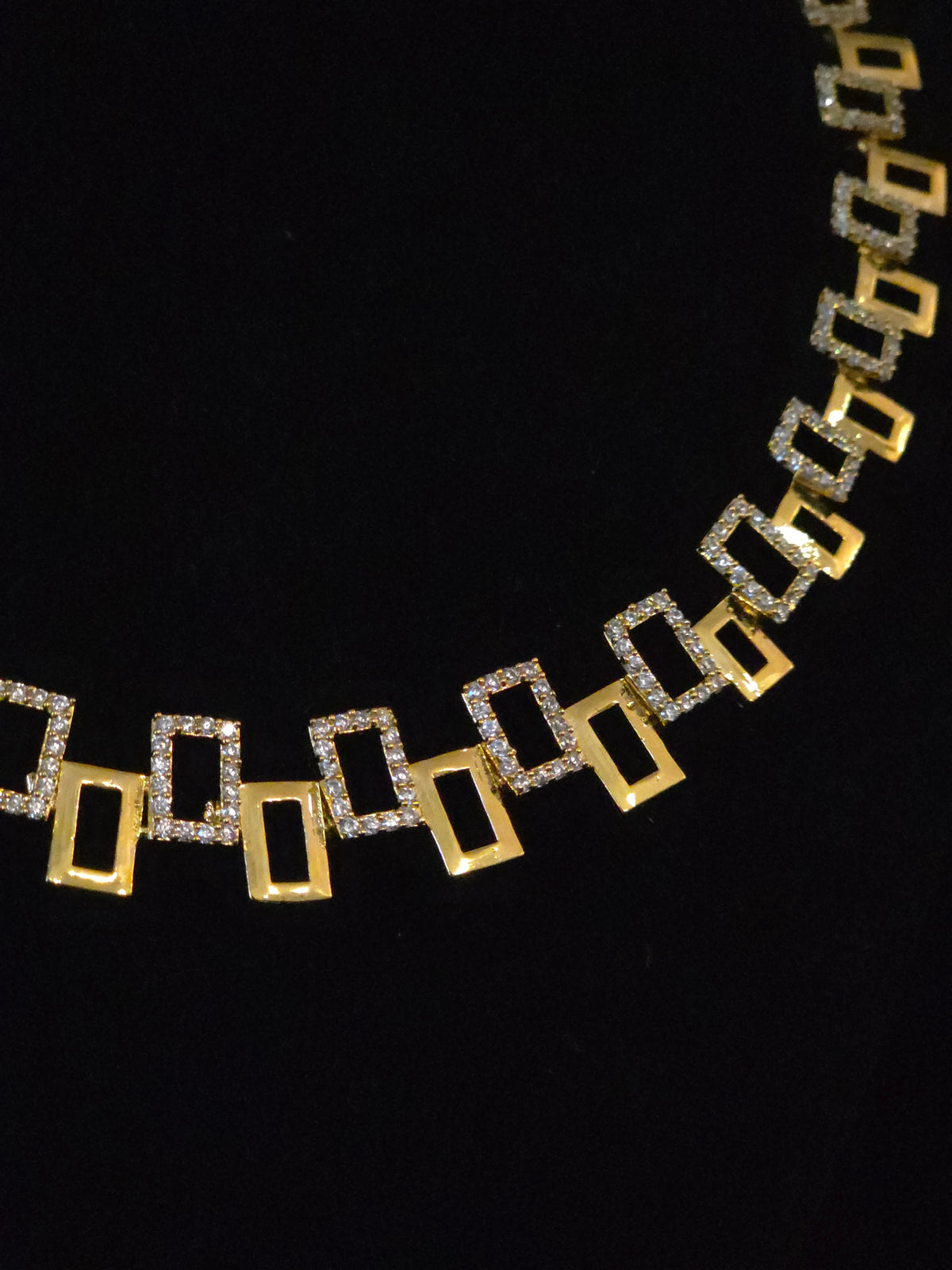 A gold geometric link necklace featuring alternating open rectangular gold links and square links paved with small clear stones, displayed against a solid black background.