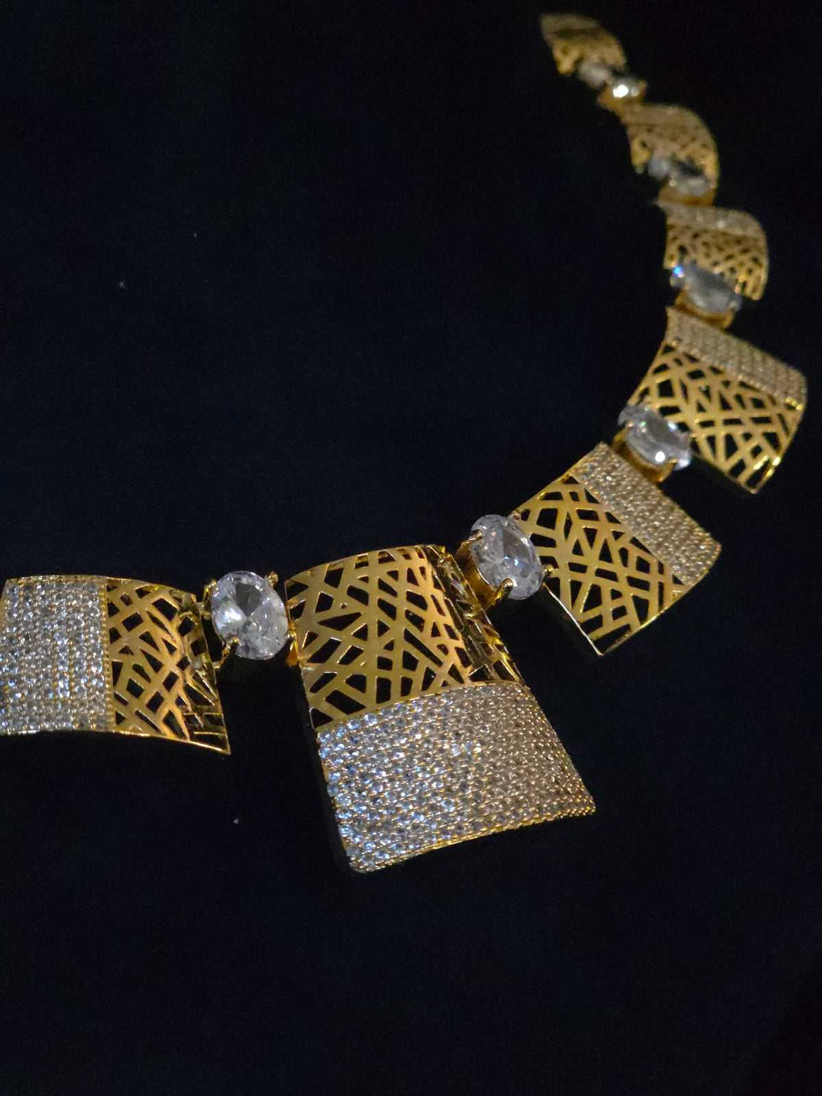A gold-toned statement necklace featuring geometric links with intricate cutout patterns, pavé-set clear stones, and a central oval clear gemstone, displayed on a black background.