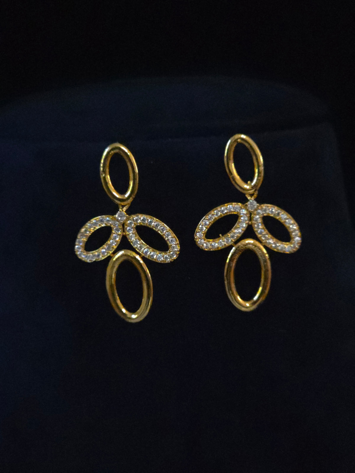 A pair of gold-tone drop earrings with oval and looped cutout details, set with small clear stones, displayed on a dark navy velvet jewelry stand.