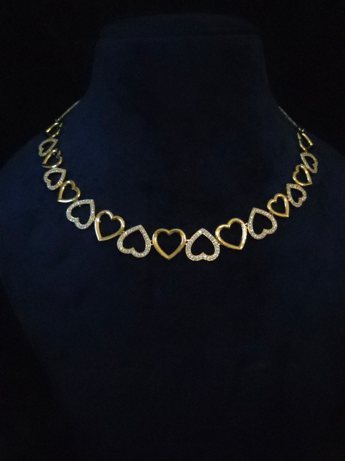 A gold heart-link necklace displayed on a black velvet jewelry bust against a black background.