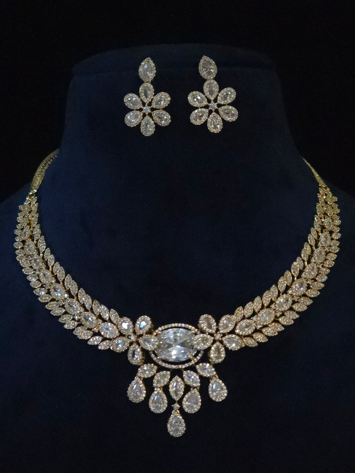 Gold-tone floral jewelry set featuring a statement necklace and matching stud earrings, embellished with clear rhinestones, displayed on a dark blue velvet neck stand against a black background.