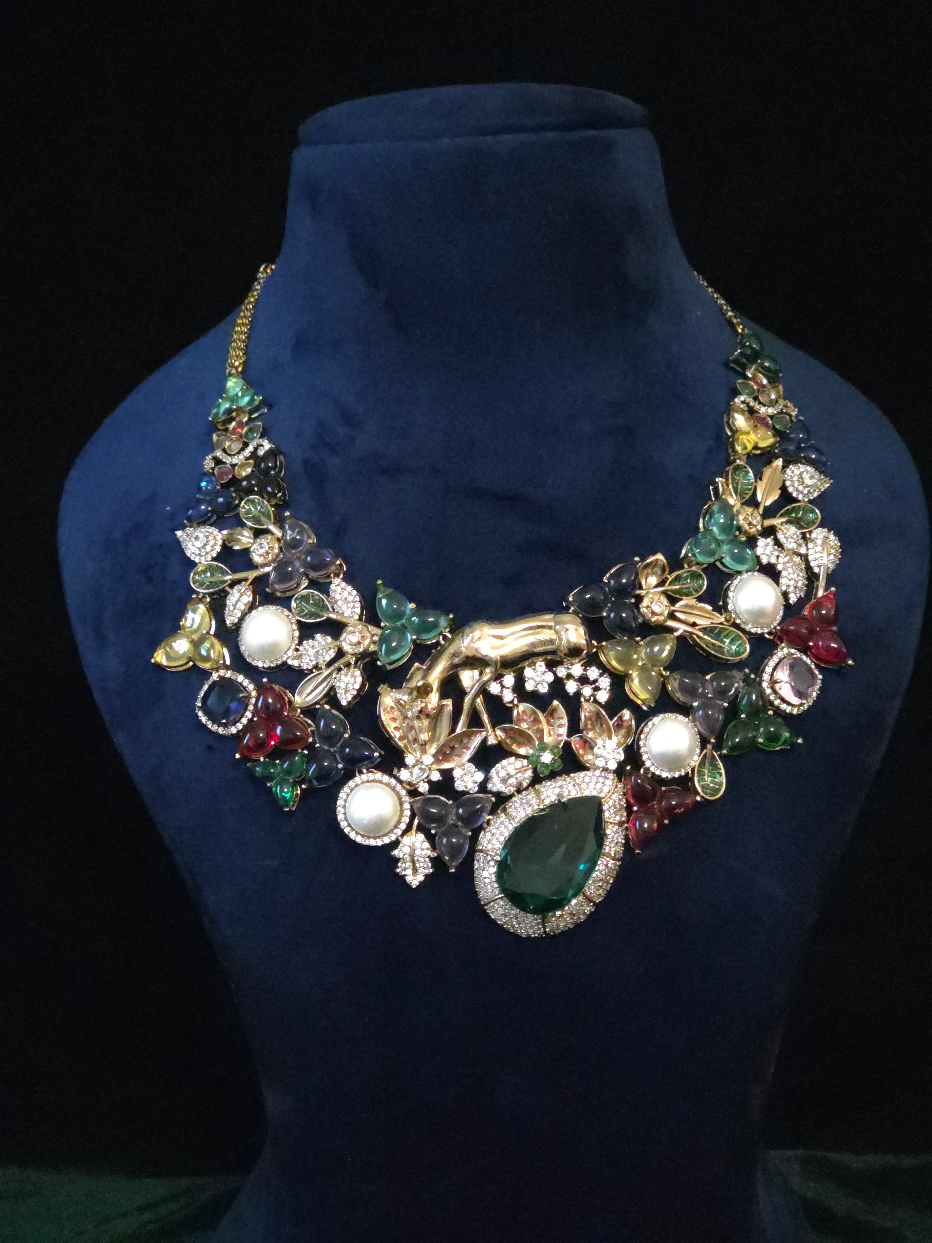 A statement bib necklace with a gold-tone animal motif and multicolored gemstones, displayed on a dark blue velvet mannequin bust against a black background.