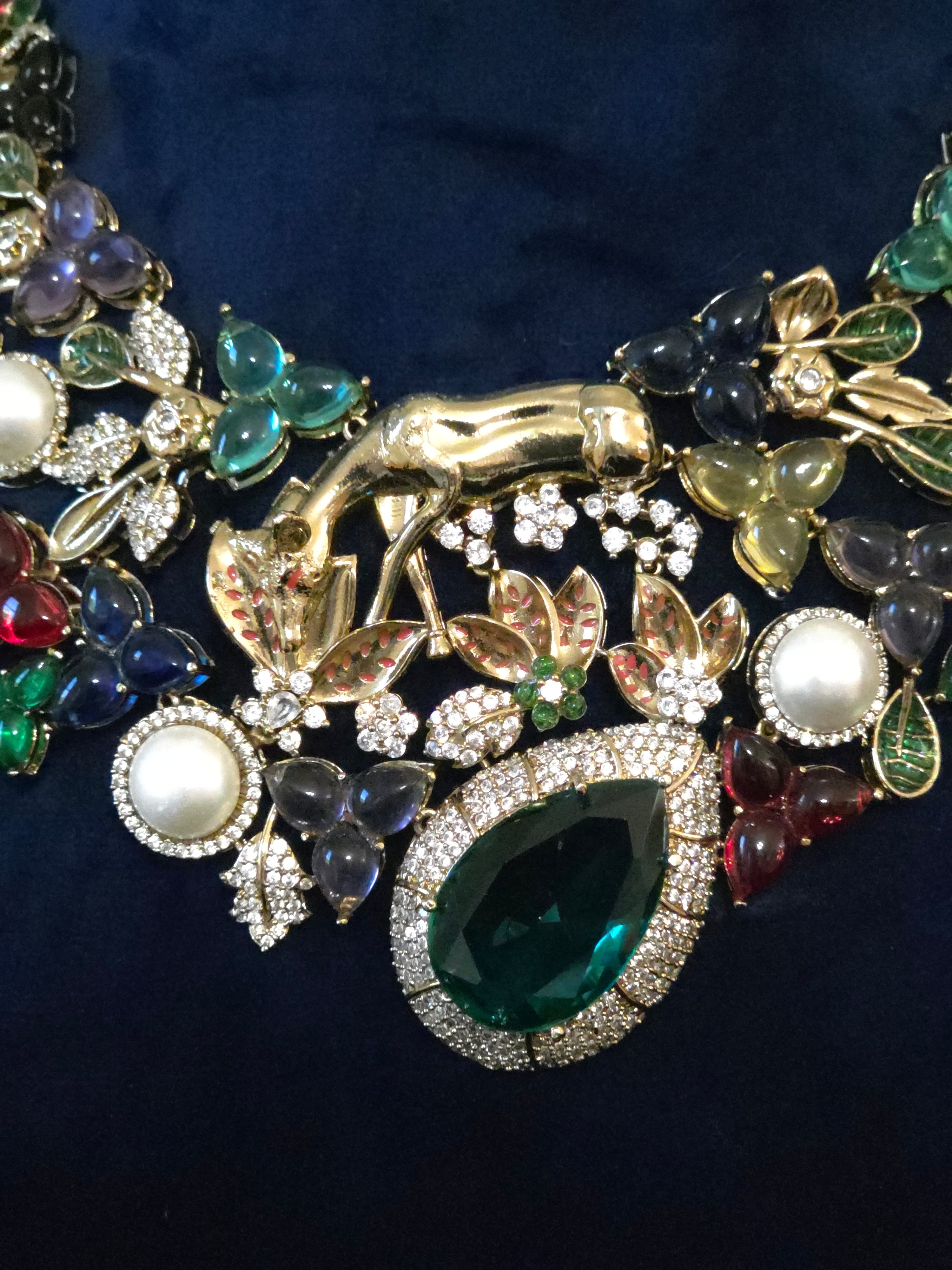 A close-up of a gold-toned statement necklace featuring a large green teardrop gemstone, white faux pearls, and multiple colorful jewel-like floral clusters on a dark navy background.