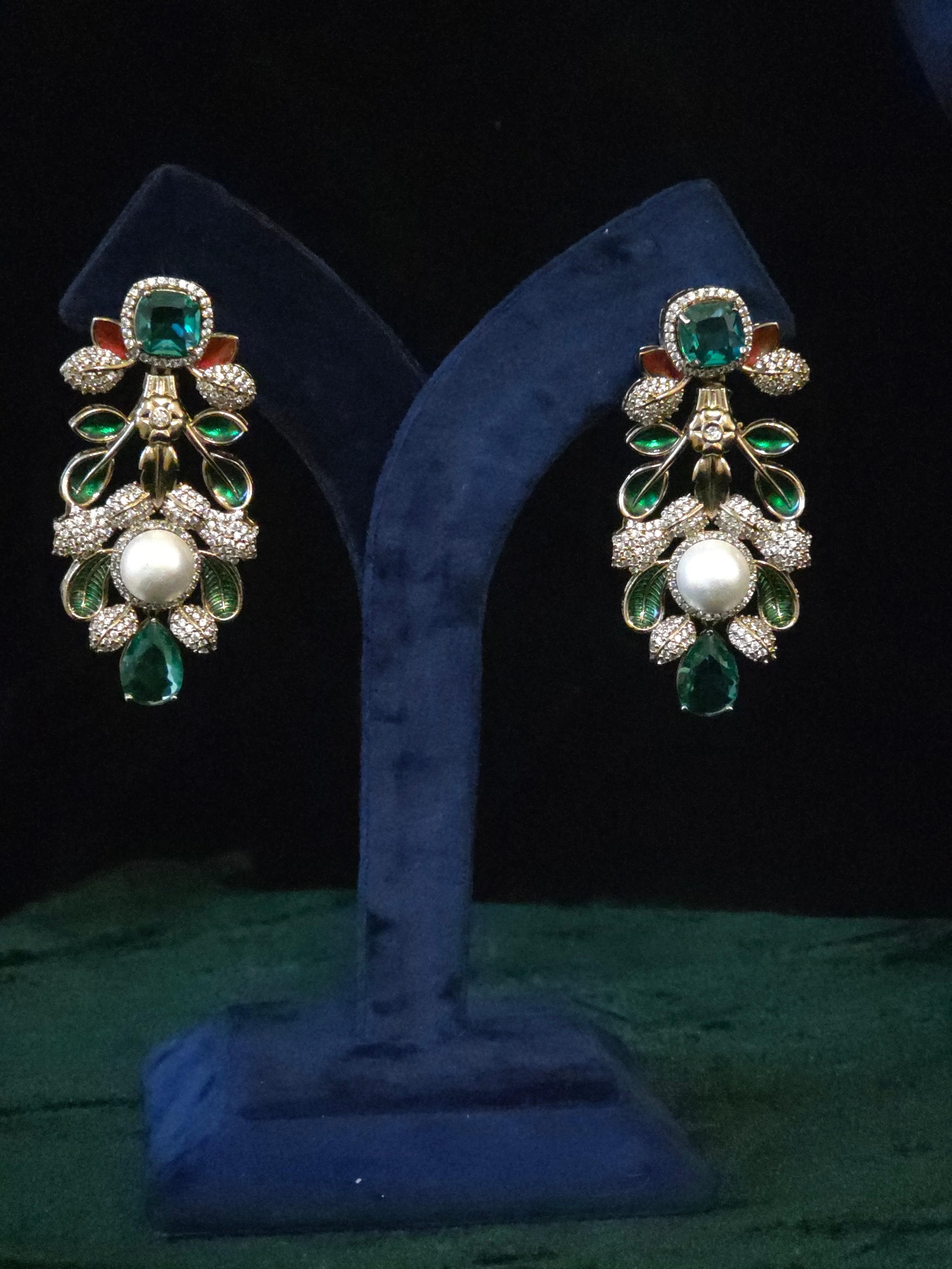 A pair of ornate drop earrings displayed on a blue velvet jewelry stand against a dark background. The earrings feature gold-toned metal settings with green teardrop and rectangular stones, a central white pearl, and small clear pavé accents.