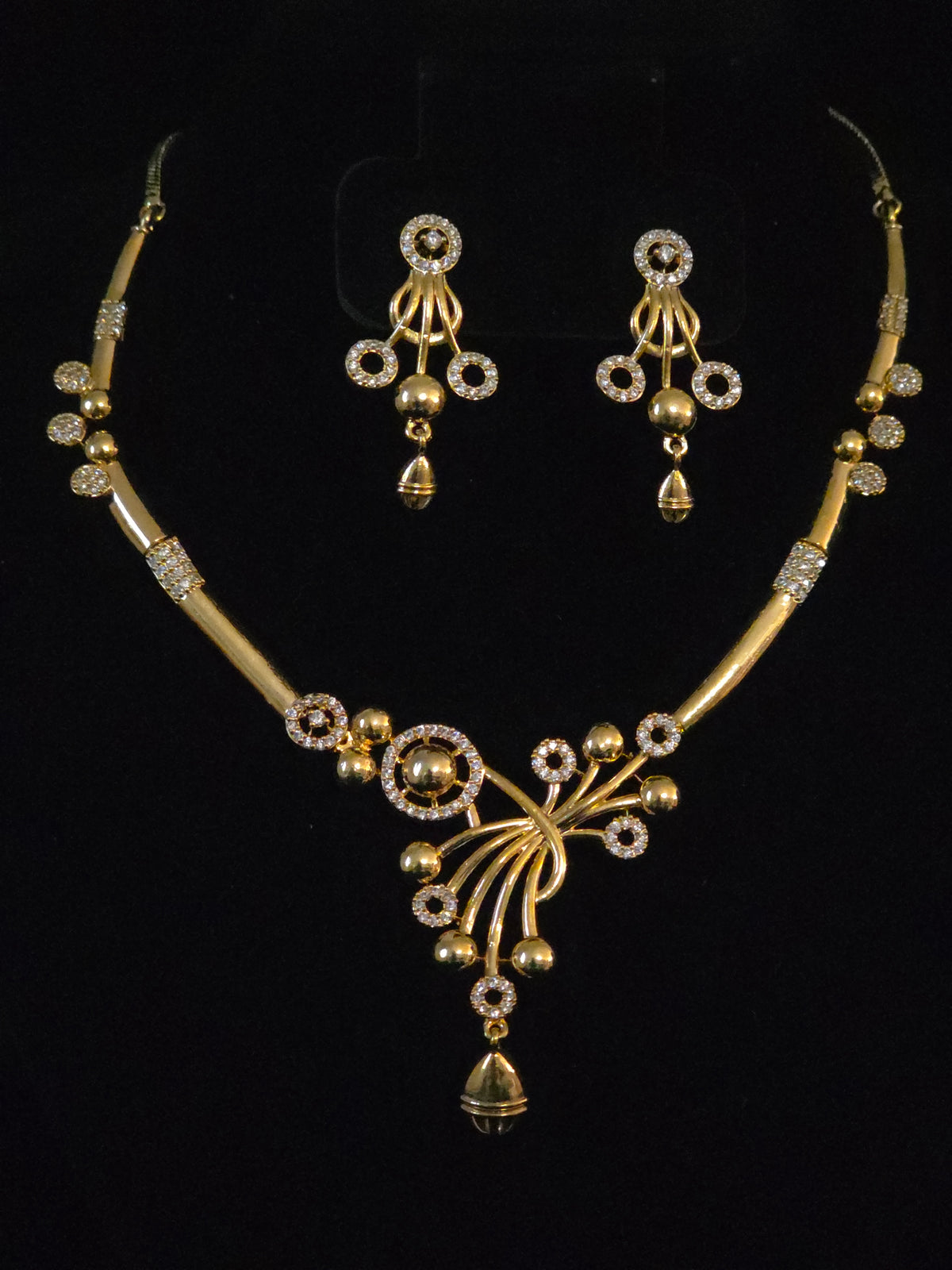 Gold-toned traditional Indian jewelry set featuring a necklace and matching earrings with intricate beadwork, floral motifs, and clear stone accents, displayed on a black background.