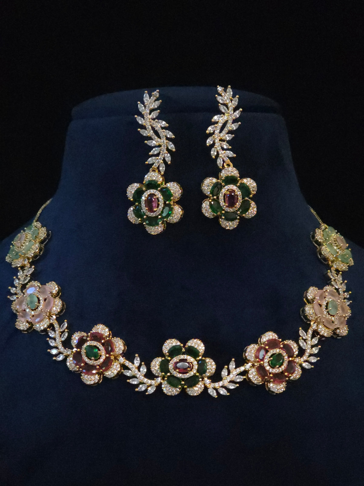 Gold floral jewelry set with a matching necklace and earrings, featuring green and pink gemstone accents, displayed on a dark blue velvet bust against a black background.