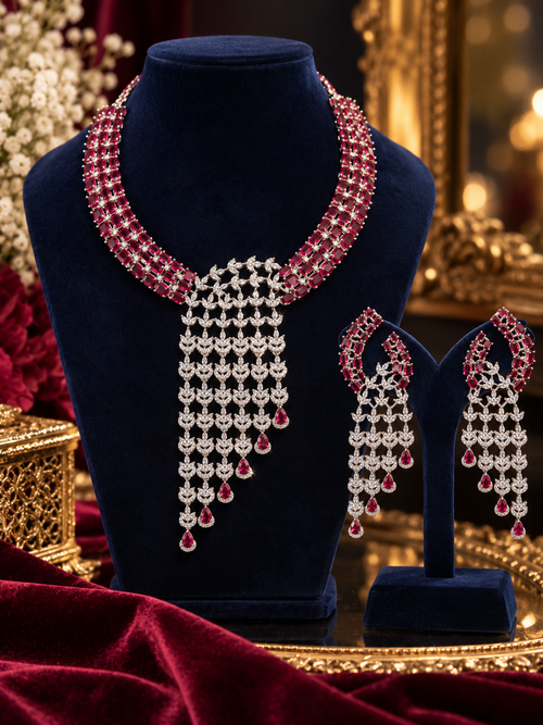 A luxury jewelry set featuring a necklace and matching earrings displayed on dark blue velvet stands. The jewelry is embellished with silver-toned metalwork and deep red gemstones, set against a rich, warm, ornate background.