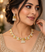 A woman wearing a gold-toned statement necklace and matching drop earrings with textured geometric details and small pearl-like accents, photographed in a softly blurred indoor setting.