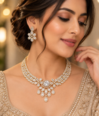 A woman wearing an ornate gold-toned jewelry set with clear stones, including a choker necklace, matching earrings, and a sleeveless embellished top. The jewelry features a central oval stone with teardrop-shaped stones below, shown against a softly blurred warm background.