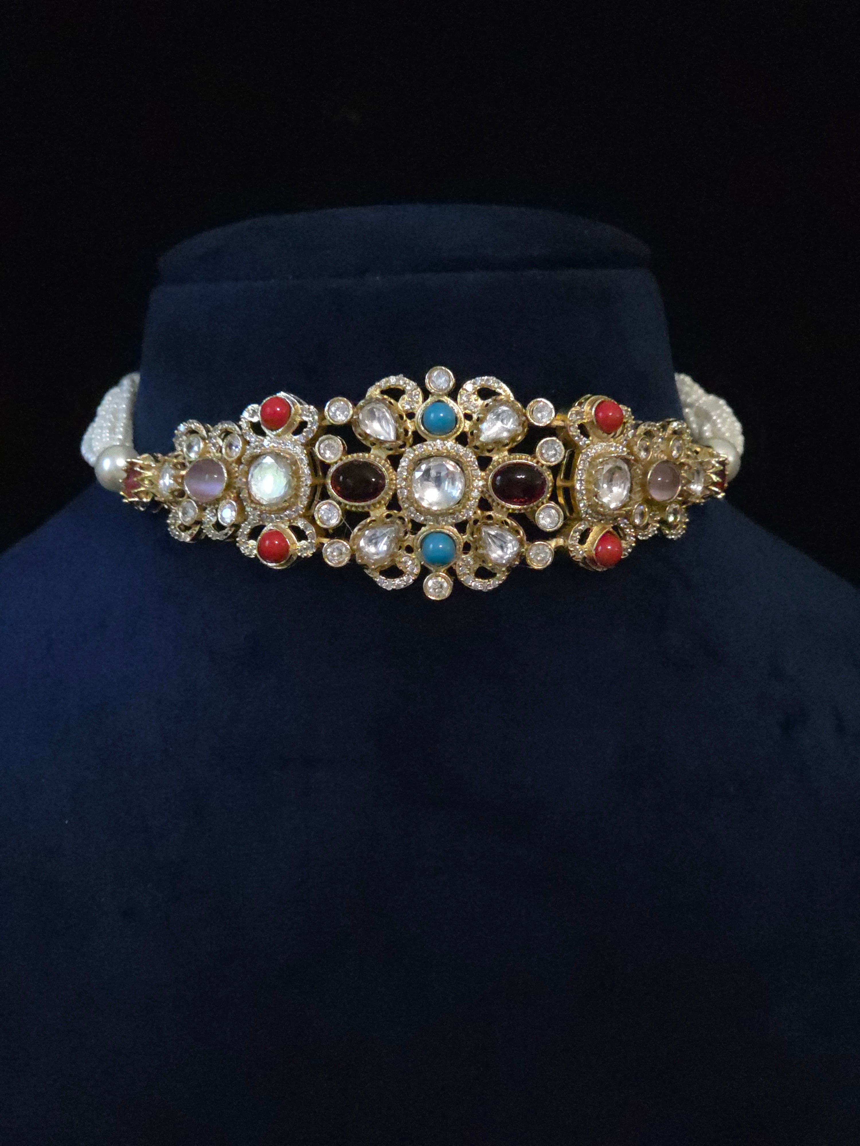 An ornate gold-toned choker necklace with multiple colorful gemstones and pearl-like accents, displayed on a dark blue velvet jewelry bust against a black background.